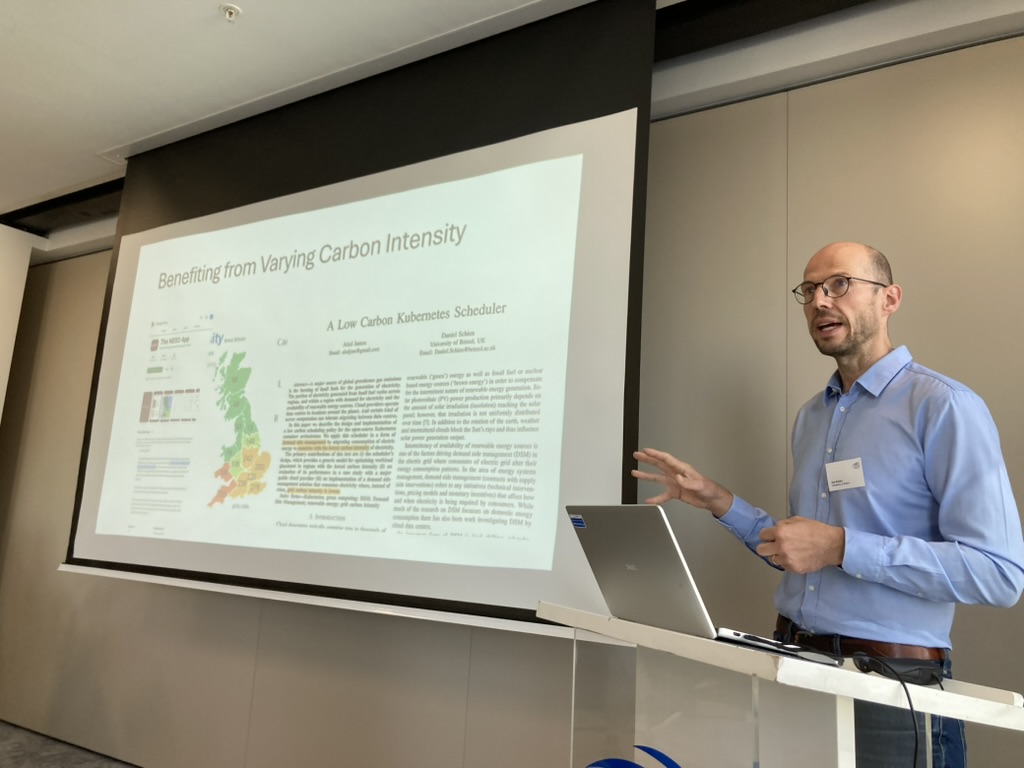 An photo of a Dr Daniel Schien, in a blue shift, presenting a slide deck in an office. The slide says "benefitting from varying carbon intensity" and shows maps of the UK, and a paper he co-authored, called "a low carbon kubernetes scheduler"